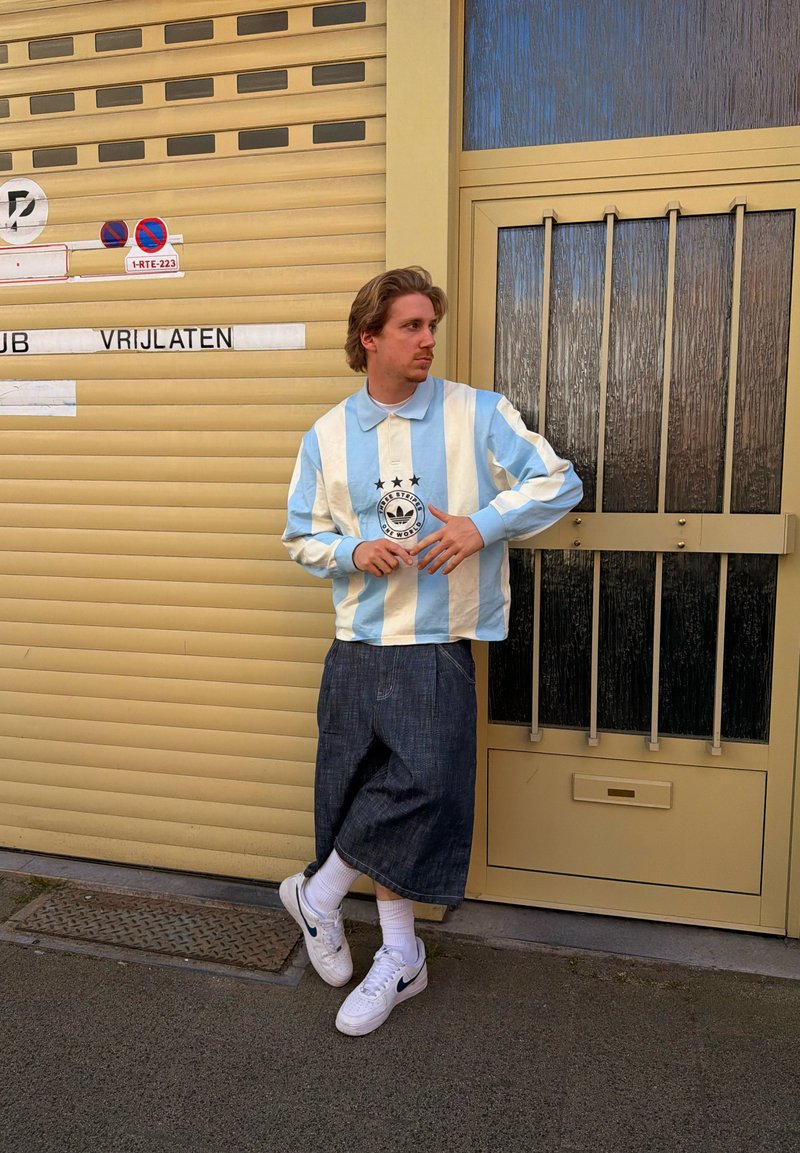 Jeune homme en chemise rayée bleu et blanc et jupe en denim s'appuie contre une porte de garage beige avec fenêtre grillagée et panneaux d'interdiction de stationner.