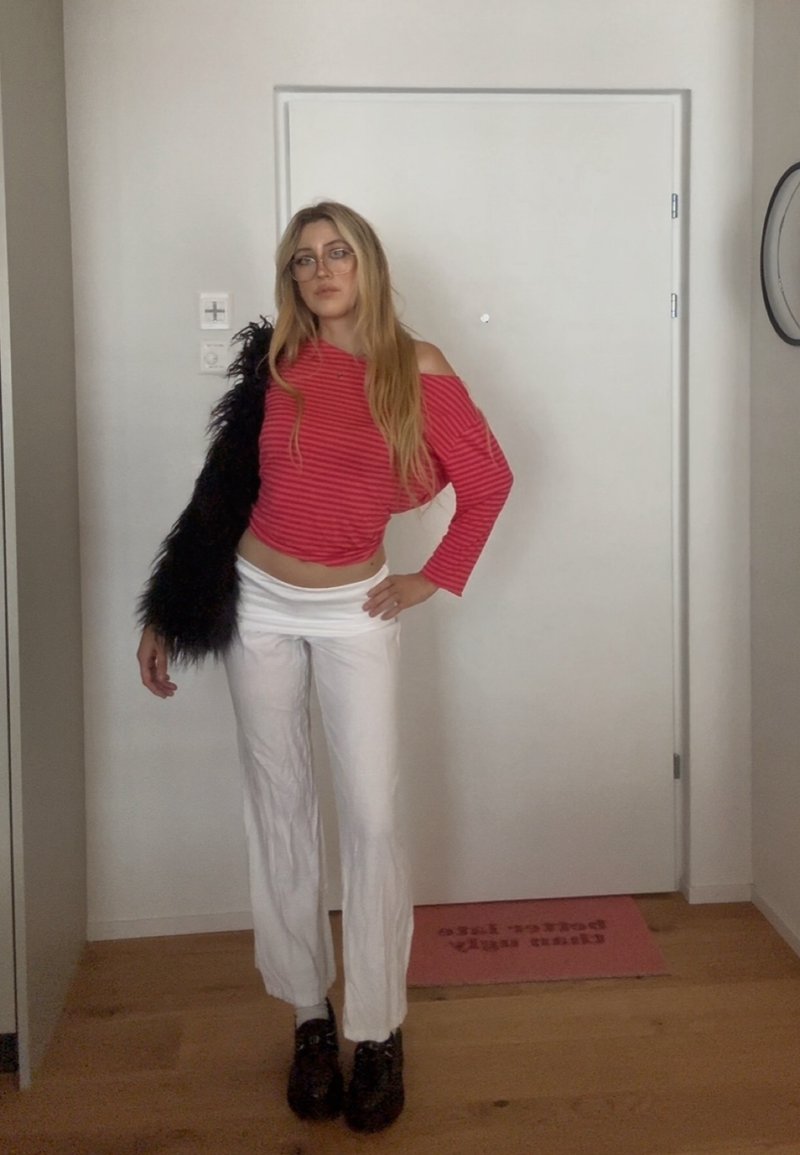 Donna con lunghi capelli biondi, che indossa occhiali, top rosso a righe con spalle scoperte, pantaloni bianchi, scarpe nere e tiene una giacca pelosa nera al chiuso.