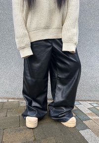 Pantaloni larghi in ecopelle nera e un maglione in maglia crema testurizzata con polsini a coste, abbinati a scarpe beige chiare con suola spessa.