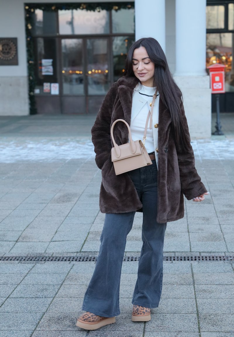 Brauner Mantel aus Kunstpelz über einem weißen Oberteil, kombiniert mit dunklen ausgestellten Jeans und beigen Plateau-Schuhen. Eine kleine beige Handtasche mit Griff oben.