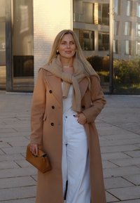 Béžový dvojradový kabát, jemná hnedá šatka, biele široké nohavice, drží malú hnedú kabelku. Hladké textúry, prispôsobený strih, mestské prostredie.