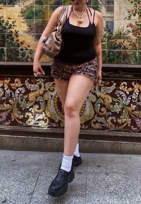 Débardeur noir, short à volants en imprimé léopard, chaussures à plateforme noires épaisses, chaussettes blanches mi-mollet, et un sac à main métallique. Fond en carreaux décoratifs.