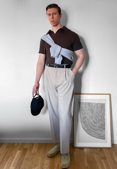 Hombre vistiendo un polo marrón oscuro, pantalones beige y zapatos claros, sosteniendo una gorra negra, con un suéter gris sobre los hombros.