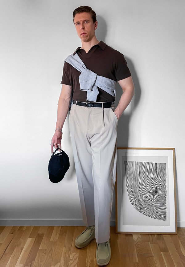 Homme portant un polo marron foncé, un pantalon beige et des chaussures claires, tenant une casquette noire, avec un pull gris drapé sur ses épaules.