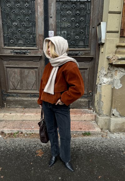 Chaqueta corta de pana marrón, bufanda beige clara, jeans negros de pierna recta y botas negras de punta, con un fondo de madera texturizada.