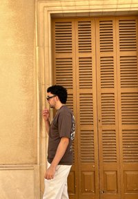 Braunes Grafik-T-Shirt, lockere weiße Hose, steht vor einer strukturierten ockerfarbenen Wand mit einer Holzjalousietür. Legere Sonnenbrille und kurze Locken.