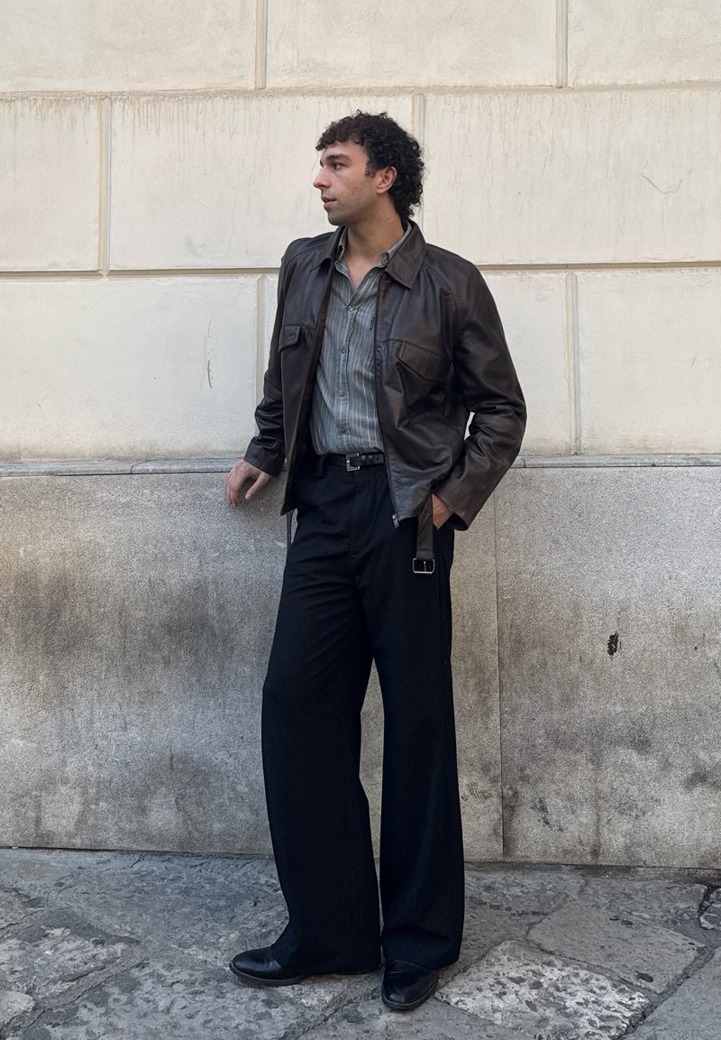 Uomo con giacca di pelle marrone, camicia a righe, pantaloni larghi neri e scarpe nere, in piedi contro un muro di pietra, che guarda verso sinistra.