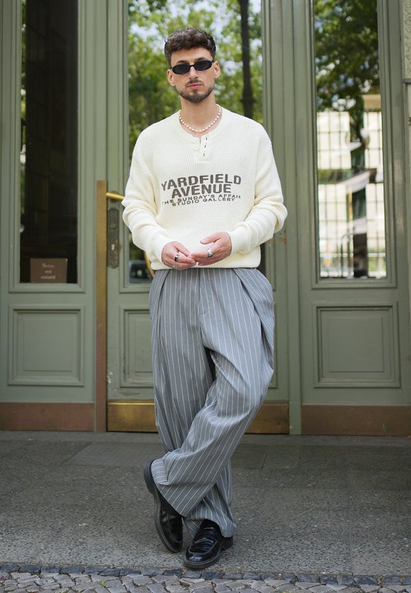 Homme portant des lunettes de soleil noires, un pull blanc texturé avec texte, un pantalon gris large à rayures fines, des chaussures noires et un collier de perles se tenant devant des portes vertes.