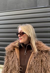 Femme aux cheveux blonds portant des lunettes de soleil foncées, un pull marron à fermeture éclair et un grand manteau marron duveteux, posant devant un fond métallique rayé.