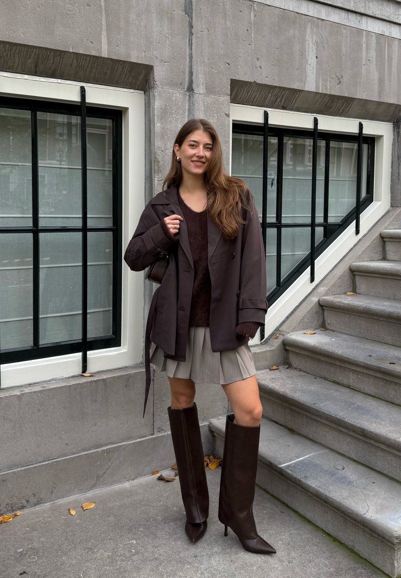 Un manteau croisé brun foncé porté sur un pull brun duveteux, associé à une jupe plissée beige et des bottes à talons aiguilles marron montant jusqu'au genou.