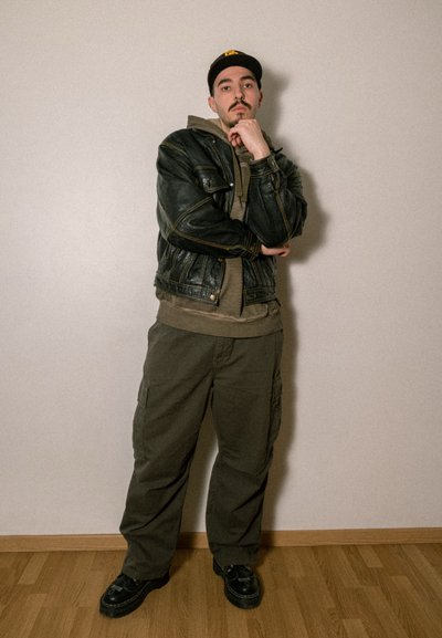 Chaqueta de cuero negra sobre una sudadera gris, combinada con pantalones cargo verde oliva y zapatos negros. El fondo es una pared neutra con suelo de madera.