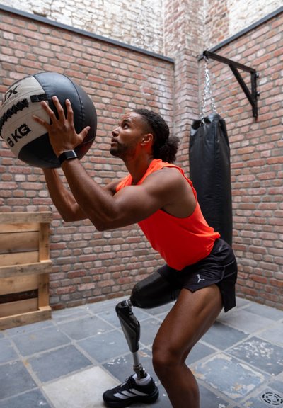 Pelota medicinal de 6 kg en blanco y negro sostenida por un hombre con una camiseta sin mangas naranja y pantalones cortos negros, con una pierna protésica, contra una pared de ladrillo.