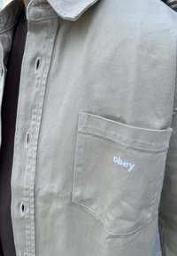 Camicia beige in corda con tasca, caratterizzata da ricamo bianco "obey". Tessuto testurizzato, design con bottoni, dettagli di cucitura visibili.