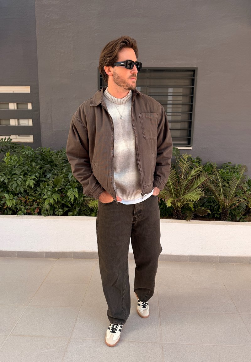 Homme portant des lunettes de soleil foncées, une veste marron, un pull clair, un pantalon foncé et des baskets blanches, debout les mains dans les poches près de plantes vertes.