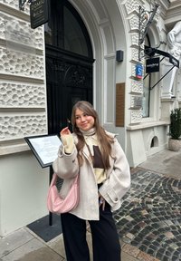 Grå yllekappa, creme- och brunsrandig tröja, svarta byxor och en rosa quiltad handväska. Kullerstengata och klassisk byggnad i bakgrunden.