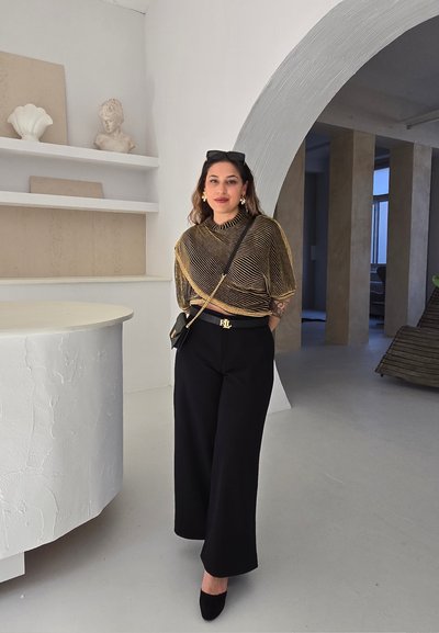 Pantalones anchos negros, blusa transparente con rayas doradas, gafas de sol negras y bolso negro pequeño. Interior minimalista con una mesa redonda y un arco.