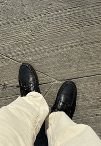 Melnas ādas kurpes ar strukturētu virsmu un auklām, kopā ar gaisā krāsotām biksēm, uz pelēka teksturēta bruģa fona.