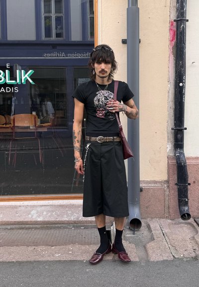 Camiseta negra con gráfico de león, pantalones cortos negros holgados, bolso burdeos y joyería de plata, combinados con calcetines y mocasines burdeos. Entorno urbano.