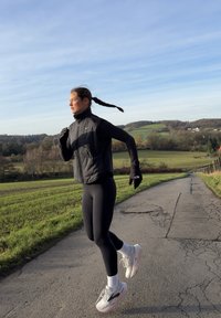 Fekete pufi mellény hosszú ujjú ing fölött, fekete leggings és fehér futócipő rózsaszín részletekkel. Fonott haj, szabadtéren egy ösvényen.