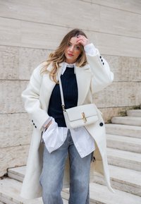 Palton alb din lână cu nasturi negri, pulover bleumarin cu model torsadat, cămașă albă și blugi bleu deschis. Poartă o geantă crem de tip crossbody cu accesorii aurii.