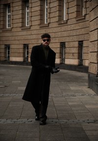 Manteau long noir, col roulé vert et gants noirs ; debout dans une rue pavée avec une architecture angulaire et peu de fenêtres en arrière-plan.