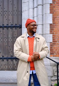Beige trench coat over a textured orange sweater and blue shirt, with a red knitted beanie and glasses, standing against a wooden and brick background.