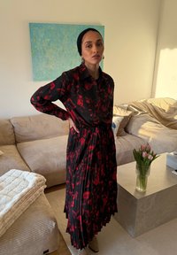 Naine, kes kannab musta ja punast lillelist kleiti ning musta pearätti, seisab elutoas, kus on beež diivan ja roosaid tulpe marmorlaual.