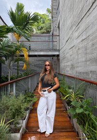 Modell i en tajt svart topp och vida vita byxor står på en trästig kantad av frodiga gröna växter och betongväggar.