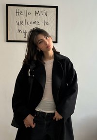 Jeune femme aux cheveux foncés portant un manteau noir et une chemise blanche, debout devant un panneau mural sur lequel est écrit « Hello MTV welcome to my ».
