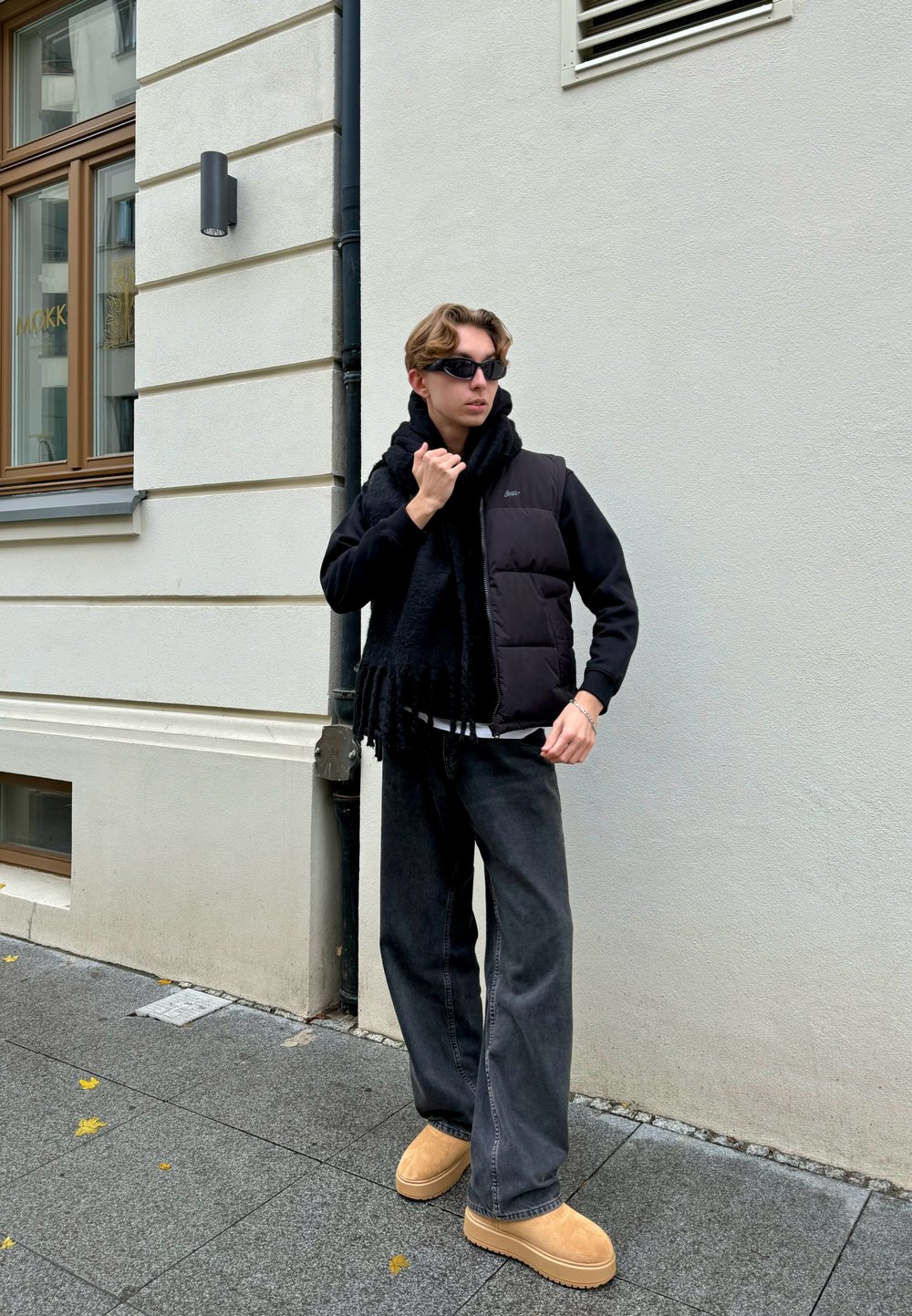 Zwarte gewatteerde vest over een zwarte hoodie, gecombineerd met wijde, donkere jeans en tan klompen. Een zwarte sjaal met franjes accentueert de outfit.
