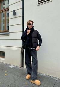 Zwarte gewatteerde vest over een zwarte hoodie, gecombineerd met wijde, donkere jeans en tan klompen. Een zwarte sjaal met franjes accentueert de outfit.