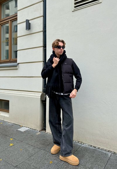 Chaleco de plumas negro sobre una sudadera con capucha negra, combinado con jeans oscuros de pierna ancha y zuecos marrones. Una bufanda negra con flecos acentúa el atuendo.