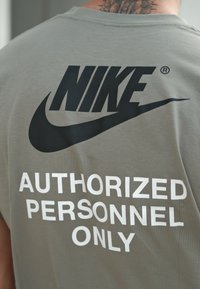 T-shirt cinza com um grande logo preto da "NIKE" e texto branco a dizer "PESSOAL AUTORIZADO APENAS" nas costas.