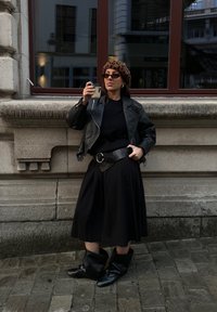 Czarna skórzana kurtka motocyklowa, czarny sweter, długa czarna spódnica, szeroki pasek z okrągłą klamrą, czarne kozaki do kolan, beret w panterkę, okulary przeciwsłoneczne.