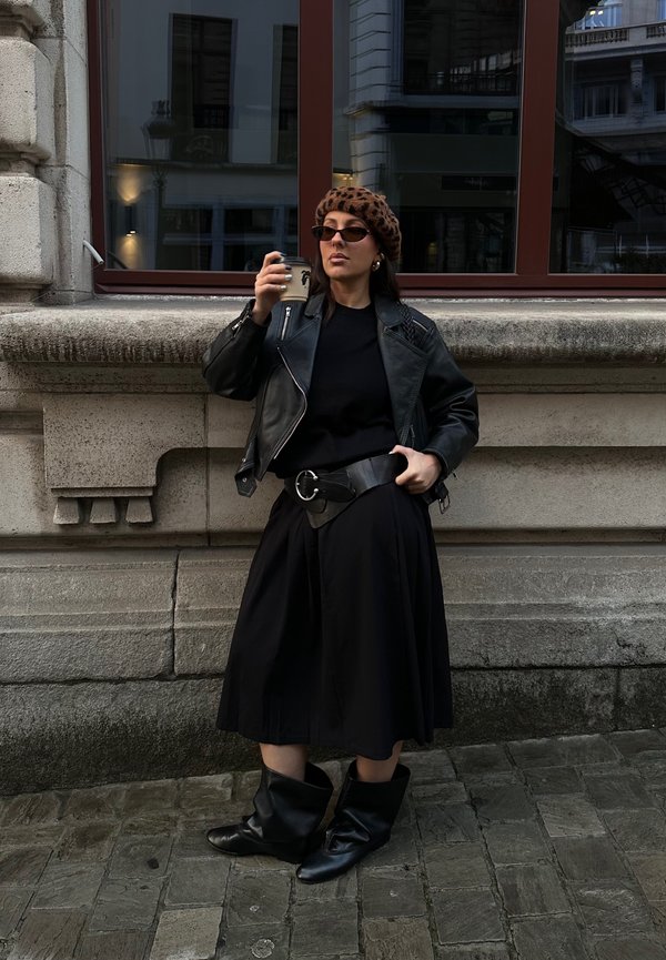 Sort lædermotorcykeljakke, sort sweater, lang sort nederdel, bredt bælte med cirkulært spænde, sorte knæhøje støvler, leopardmønstret baret, solbriller.