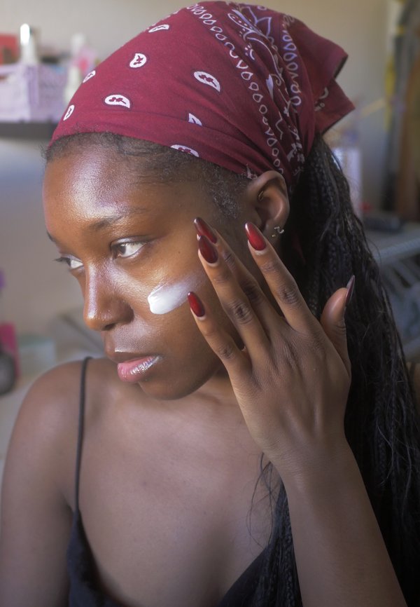 Modell som påfører en hvit krem på kinnet, iført et burgunderfarget bandana og svarte klær. Lange røde negler fremhever hånden hennes.
