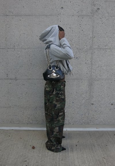 Sudadera con capucha gris, pantalones cargo de camuflaje y un bolso negro con una correa de cadena plateada y acentos metálicos, contra una pared de concreto.