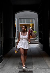 Donna in abito bianco e sandali che passeggia attraverso un arco scuro verso un moderno negozio con una vetrina in mostra.