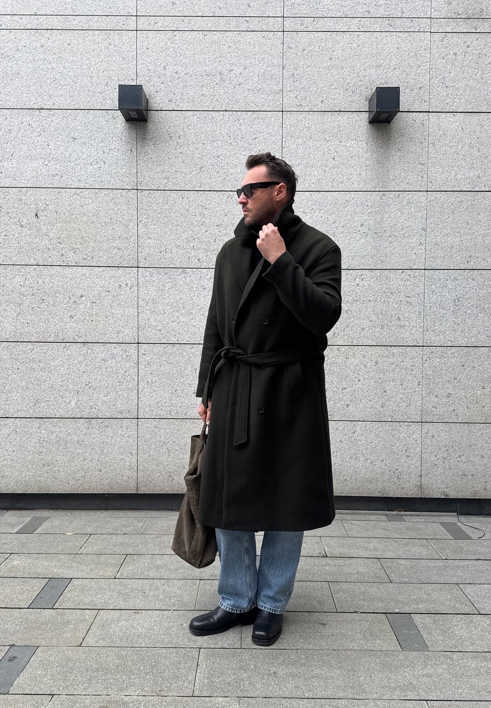 Homme portant un long manteau noir et des lunettes de soleil, tenant un sac fourre-tout marron, debout contre un mur carrelé gris clair avec deux appliques murales noires.
