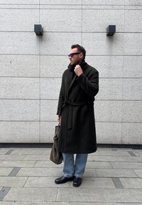 Homme portant un long manteau noir et des lunettes de soleil, tenant un sac fourre-tout marron, debout contre un mur carrelé gris clair avec deux appliques murales noires.