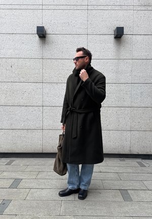 Hombre con abrigo largo negro y gafas de sol sosteniendo un bolso tote marrón, de pie frente a una pared azulejada de color gris claro con dos luces de pared negras.