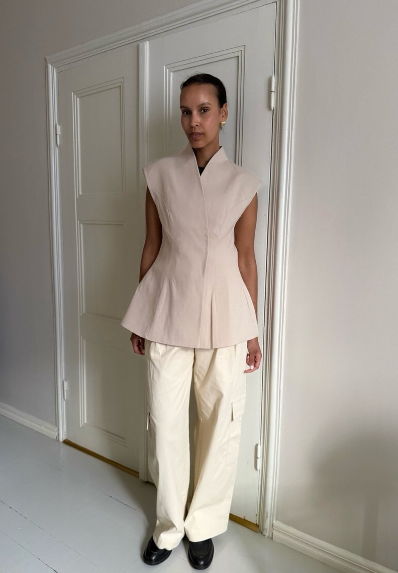 Femme debout à l'intérieur portant un haut beige sans manches sur mesure, un pantalon cargo ample couleur crème et des chaussures noires devant une porte blanche.