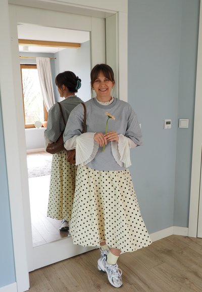 Joven mujer sonriendo, sosteniendo una flor, vistiendo un suéter a rayas, blusa blanca con mangas acampanadas, falda con lunares, zapatillas deportivas, de pie junto a un espejo en el interior.