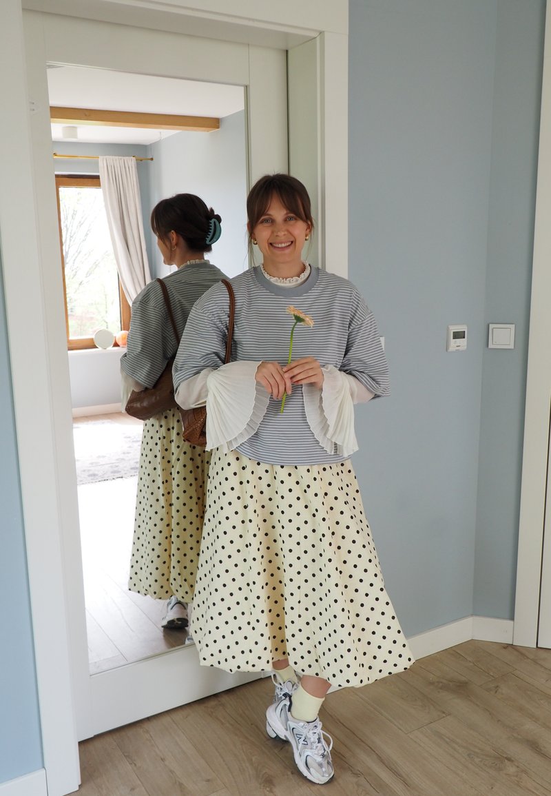 Jeune femme souriante, tenant une fleur, portant un pull rayé, une blouse blanche à manches évasées, une jupe à pois, des baskets, debout près d'un miroir à l'intérieur.