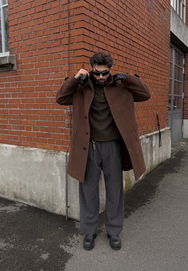 Muž v hnědém kabátě, černých rukavicích, slunečních brýlích, šedých kalhotách a černých botách stojí u cihlové budovy a upravuje si límec kabátu.