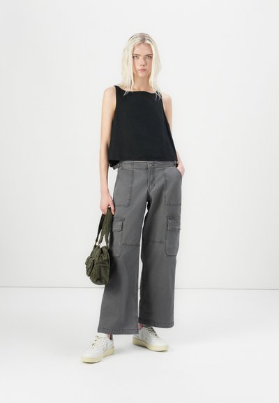 Top negro sin mangas con escote redondeado, combinado con pantalones cargo grises de pierna ancha que cuentan con bolsillos laterales. Zapatillas blancas y una bolsa verde.