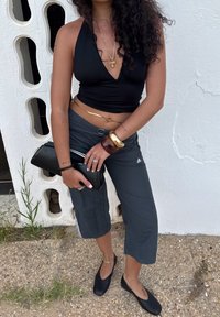 Top negro con cuello halter, pantalones cropped grises con rayas laterales, bolso de mano negro, pulsera dorada y zapatos planos negros. Fondo texturizado.