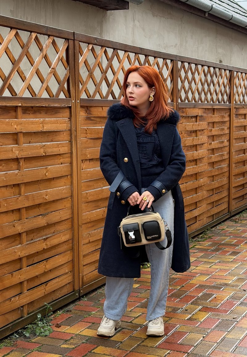 Femme aux cheveux roux portant un manteau noir et tenant un sac à main beige et noir, debout sur un chemin en briques mouillé à côté d'une clôture en bois.
