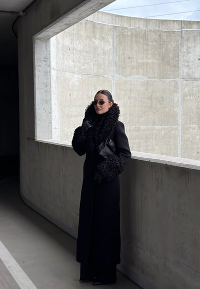 Abrigo negro de lana con un cuello y puños voluminosos y texturizados, combinado con guantes negros y gafas de sol, sobre un fondo de pared de hormigón.