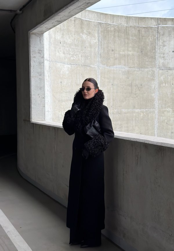 Abrigo negro de lana con un cuello y puños voluminosos y texturizados, combinado con guantes negros y gafas de sol, sobre un fondo de pared de hormigón.